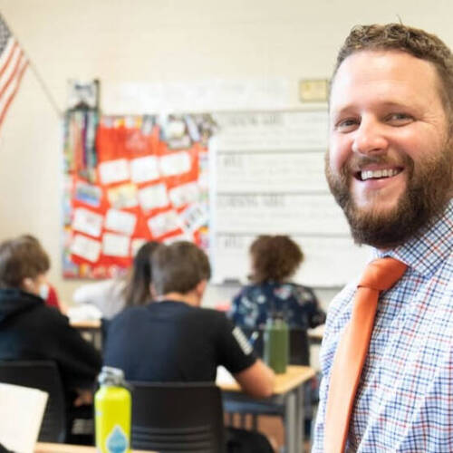 A smiling person in a classroom, wearing a checkered shirt and an orange tie, with students seated in the background.
